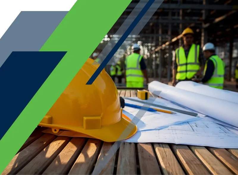 A yellow hard hat and rolled construction plans on a wooden table, with workers in high-visibility vests behind